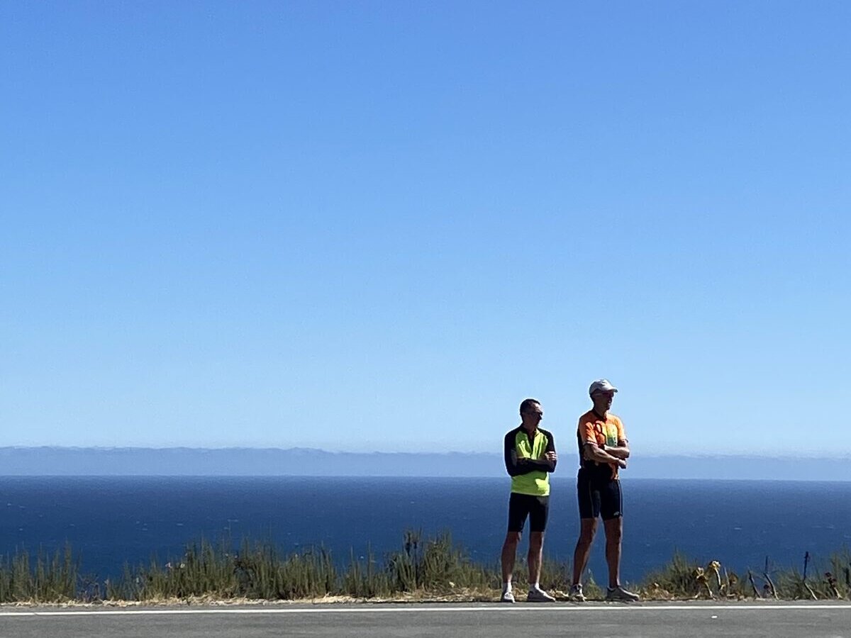 Two cyclists in bright jerseys standing with arms crossed at the road's edge, gazing at the water