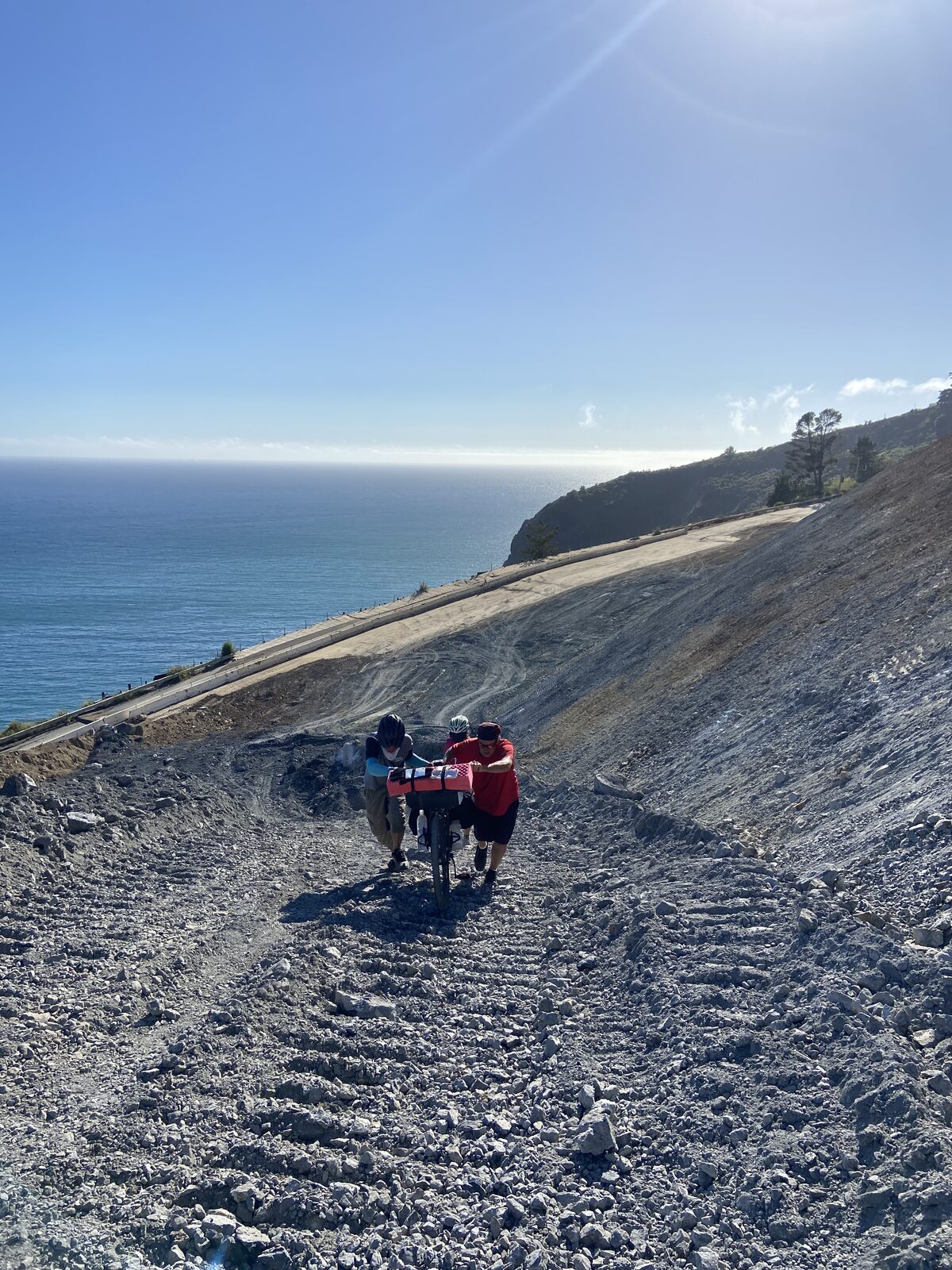 Two cyclists pushing a loaded bicycle up a steep gravel landslide above the ocean