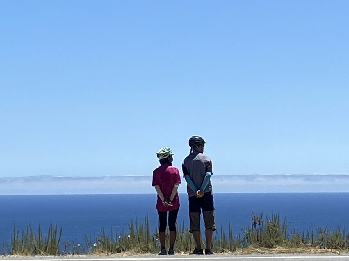 A couple standing side by side, hands clasped behind their backs, both facing the ocean