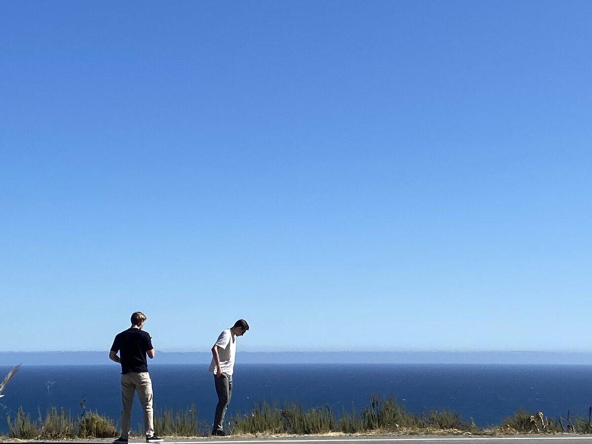 Two men in dark and white shirts standing at the cliff edge, looking out over the ocean
