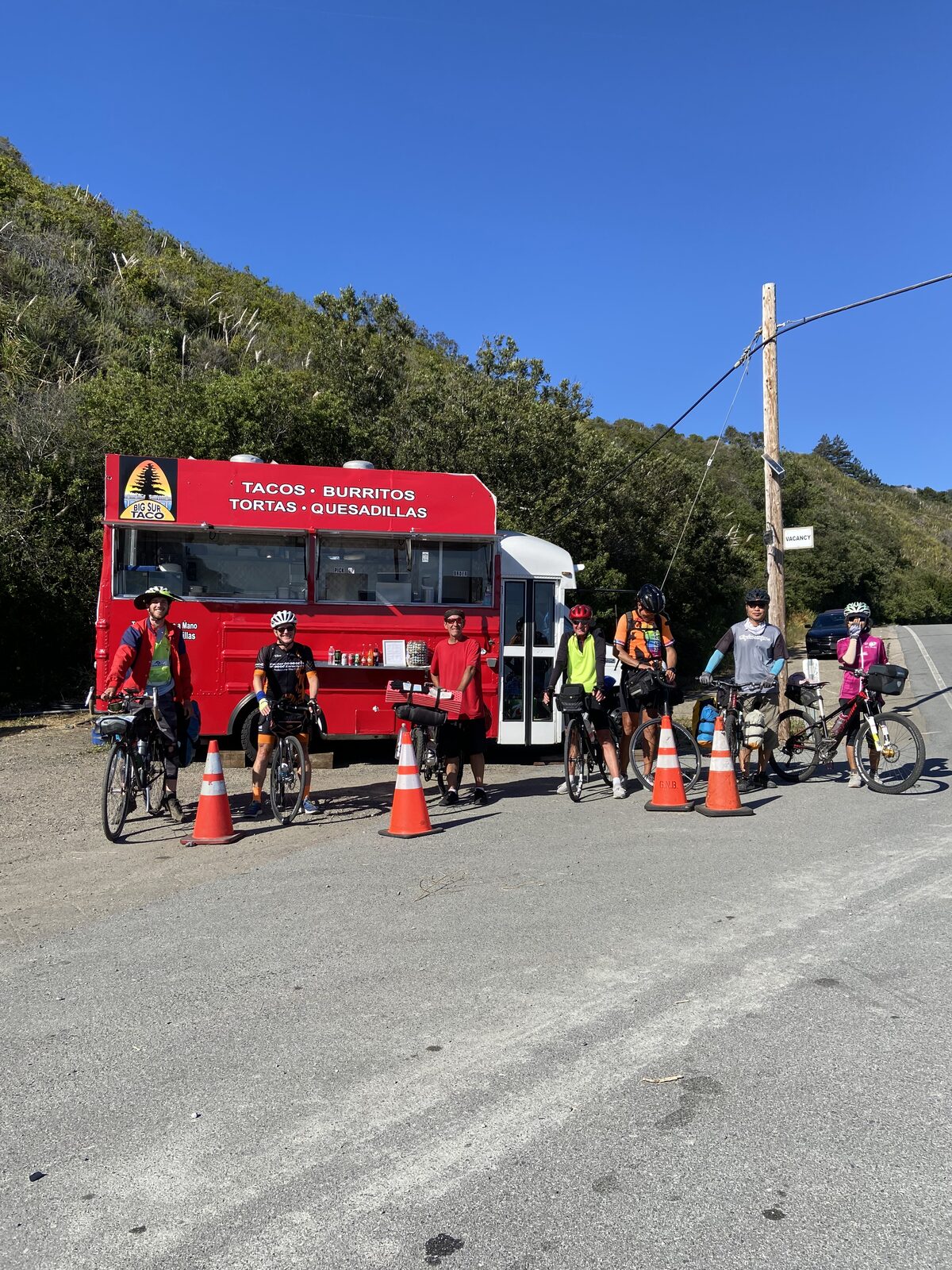 A group of cyclists with loaded bikes lined up in front of the bright red Big Sur Taco Truck, hillside behind them